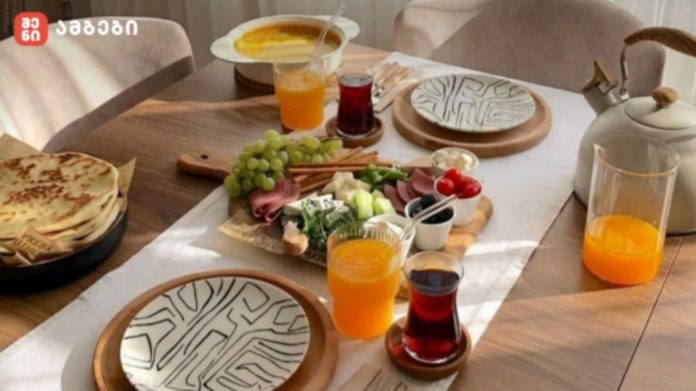 Breakfast spread on a wooden table: flatbreads, a cheese and charcuterie board with grapes, greens, olives, and cherry tomatoes, plus orange juice and tea/coffee beverages with a teapot nearby.