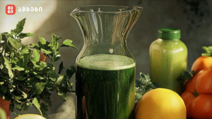 Green juice in a glass pitcher on a kitchen counter, surrounded by leafy greens and citrus fruits like a lemon and oranges nearby.
