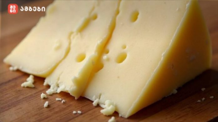 Wedge of pale yellow Swiss cheese with holes on a wooden cutting board, with a small logo in the top-left corner.