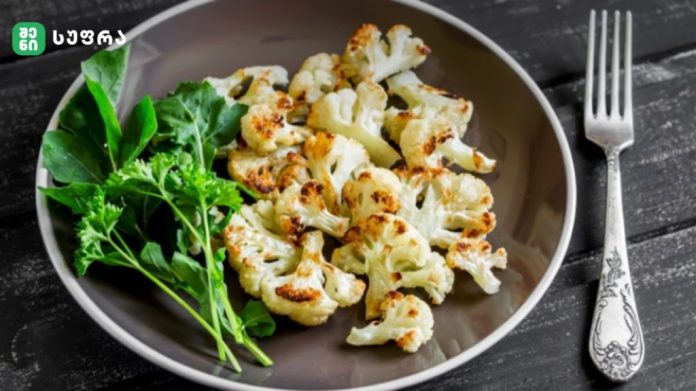 Roasted cauliflower florets on a gray plate with fresh greens on a dark wooden table, with a silver fork nearby.