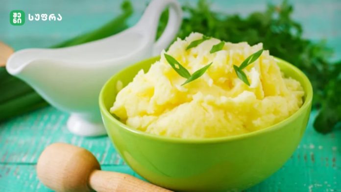 Green bowl of mashed potatoes garnished with green chive pieces, with a white gravy boat in the background.