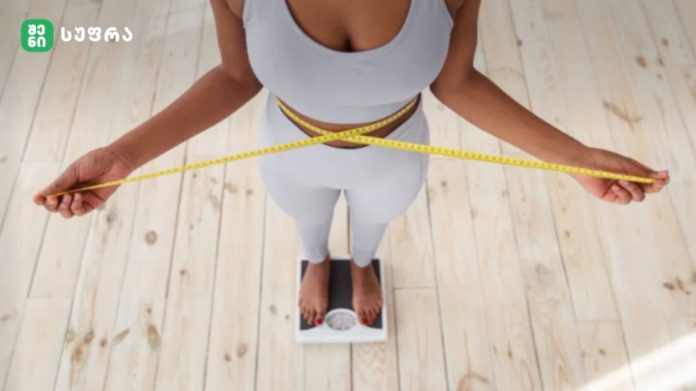 Top-down view of a person standing barefoot on a scale, wrapping a yellow measuring tape around their waist.