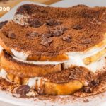 Stacked chocolate-topped dessert with cream filling, cocoa powder, and chocolate chunks on a white plate (logo watermark in the top-left).