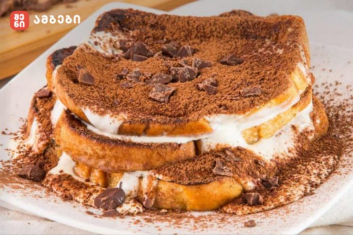 Stacked chocolate-topped dessert with cream filling, cocoa powder, and chocolate chunks on a white plate (logo watermark in the top-left).
