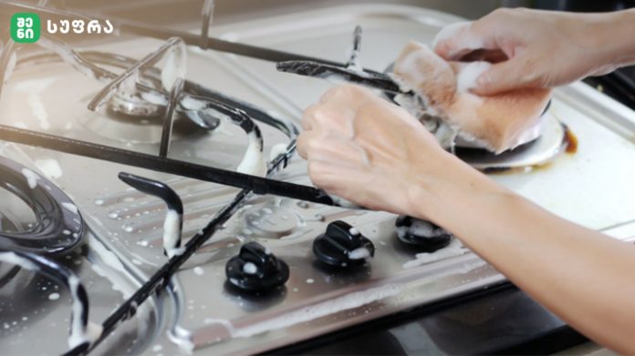Person scrubbing a dish with soapy water on a stainless steel kitchen sink area, glasses and utensils nearby