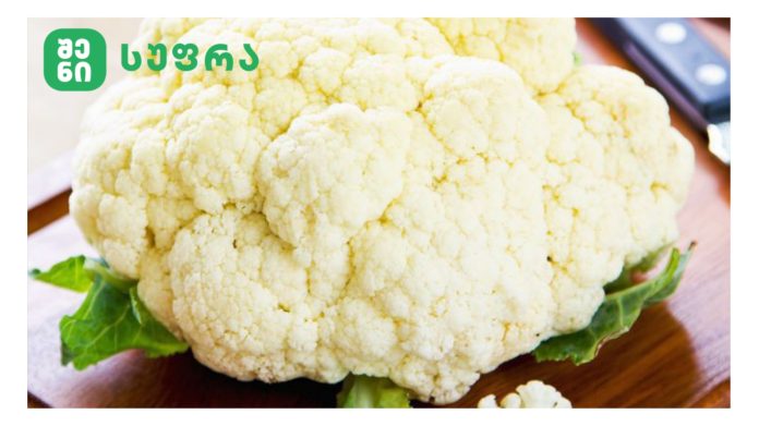 Yvavilovani-kombosto Close-up of a large cauliflower head resting on green leaves on a wooden cutting board, with a knife in the background.
