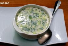 Აკროშკა – ცივი წვნიანი ზაფხულში Creamy green herb soup in a white bowl on a pale plate with a spoon beside it; garnished with chopped greens.