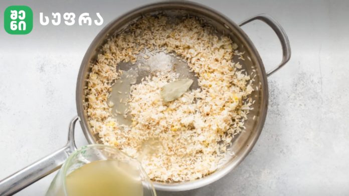Metal pot of rice with a bay leaf, broth being poured in from a pitcher nearby.