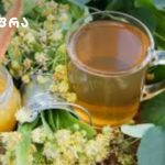 Glass teapot and cup filled with amber tea, set among yellow flowers and green leaves (branding logo visible in top-left).