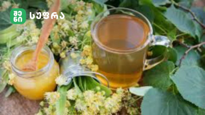 Glass teapot and cup filled with amber tea, set among yellow flowers and green leaves (branding logo visible in top-left).