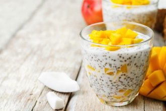 Chia pudding with yellow mango cubes in a clear glass on a rustic wooden table with coconut pieces nearby.