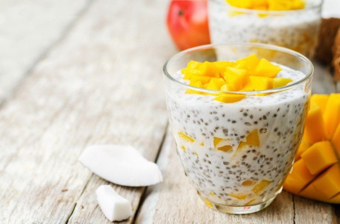 Chia pudding with yellow mango cubes in a clear glass on a rustic wooden table with coconut pieces nearby.