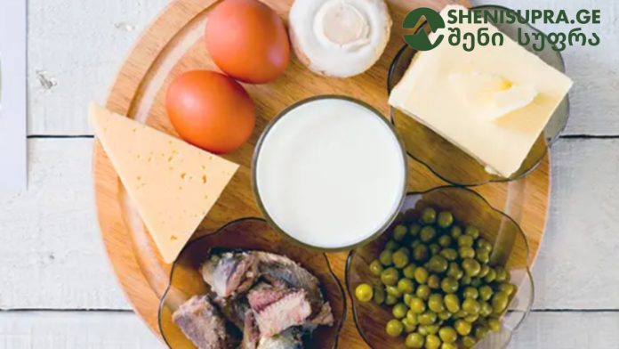 Dairy and deli spread on a wooden board: cheese wedges, butter, eggs, a glass of milk, peas, and fish pieces with a logo in the corner