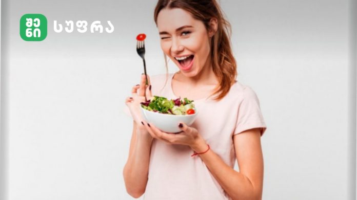 Smiling woman in a pale pink shirt holding a bowl of salad, winking with a fork holding a cherry tomato, against a light gray background (logo in top-left).