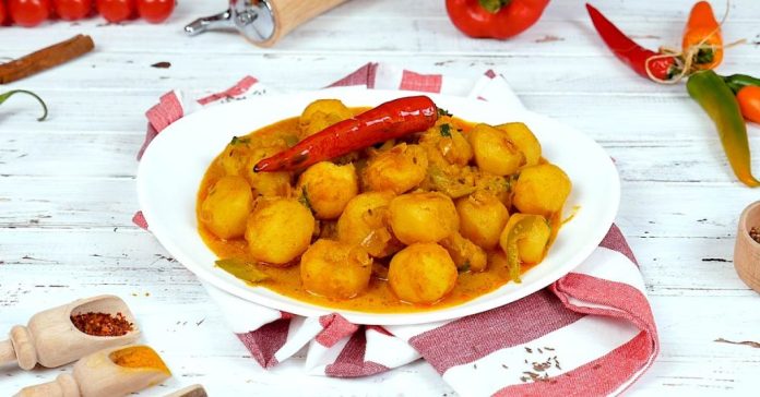 White bowl of spicy potato curry with a whole red chili on top, on a white wooden table.