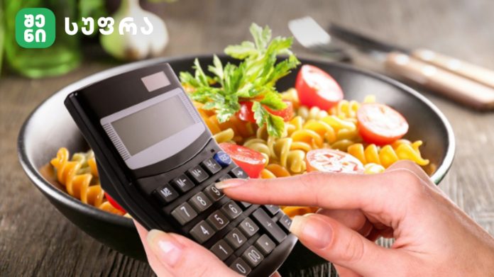 Person holding a handheld calculator over a bowl of pasta with cherry tomatoes and parsley in a kitchen setting