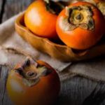 Three bright orange persimmons in a brown paper bag on a rustic wooden table, with a dark backdrop