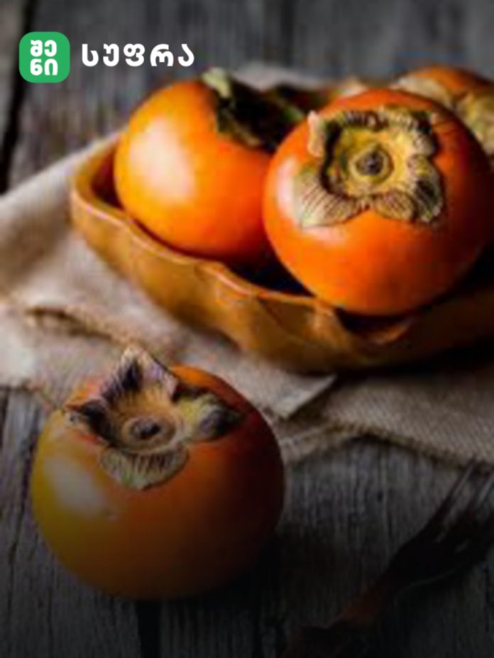 Three bright orange persimmons in a brown paper bag on a rustic wooden table, with a dark backdrop