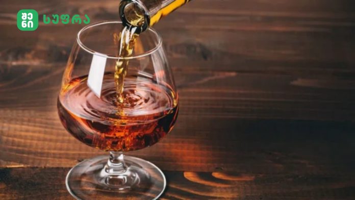 Pouring red wine into a stemmed glass on a dark wooden table, with a green logo in the top-left corner.