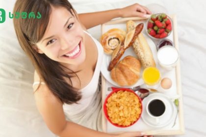 Smiling woman in bed with a tray of breakfast: croissants, pastries, cereal, fruit, eggs, juice, milk, and coffee.