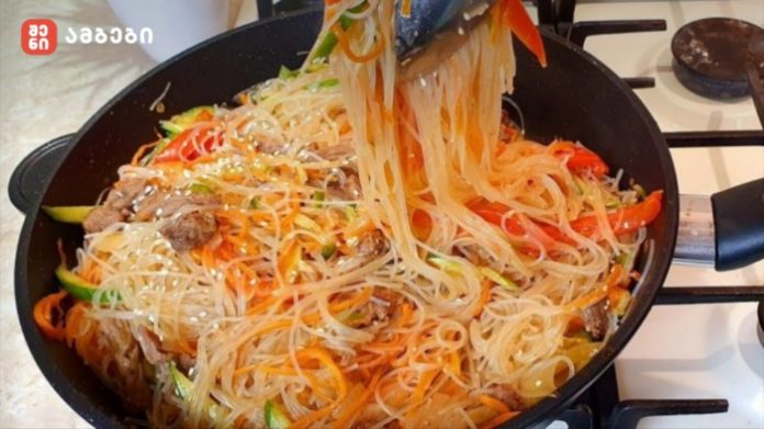 Stir-fried glass noodles with beef and colorful vegetables in a black skillet on a gas stove.