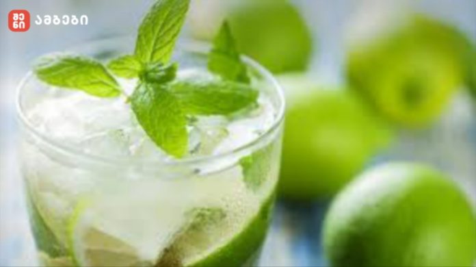 Glass of mojito with fresh mint leaves and lime wedges, ice cubes, blurred green limes in the background on a bright surface