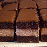 Brownie squares with a dense chocolate base, creamy middle layer, and glossy chocolate top on a plate.
