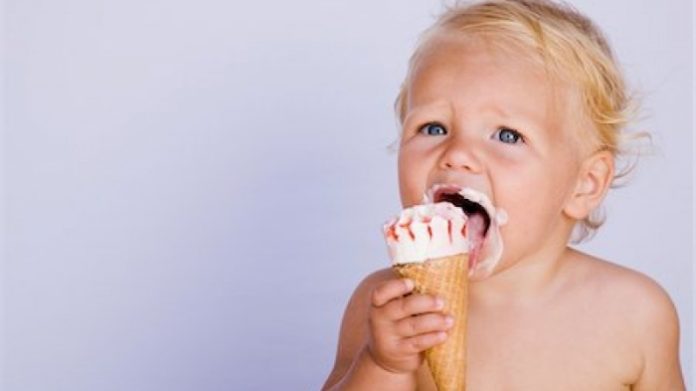 Young toddler with blond hair licking a vanilla ice cream cone with red strawberry sauce on top.