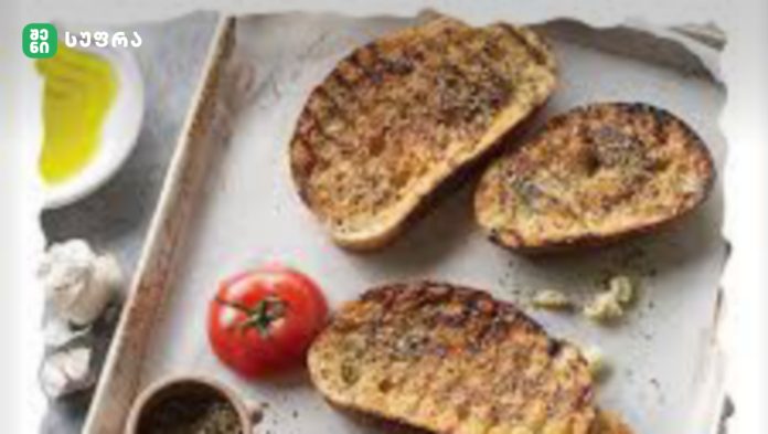 Slices of toasted bread topped with seeds and herbs on a parchment-lined baking tray, with a tomato and dipping bowls nearby.
