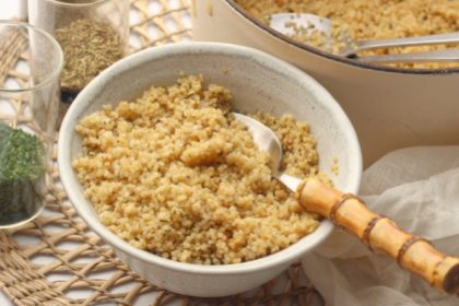 Bowl of fluffy couscous with a spoon, on a woven placemat with herbs and a pot in the background.