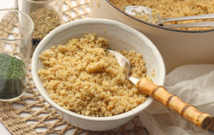 Bowl of fluffy couscous with a spoon, on a woven placemat with herbs and a pot in the background.