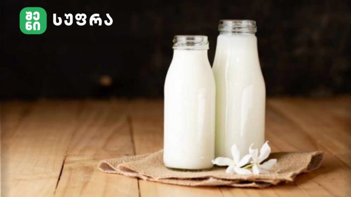 Two white milk bottles on a wooden table with a beige cloth and white flowers nearby.