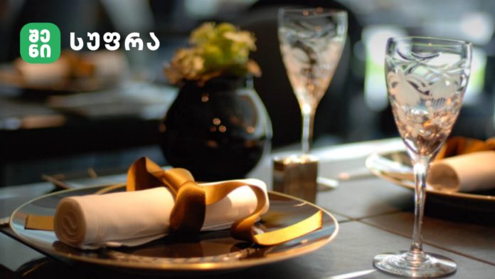 Elegant table setting: rolled white napkin tied with a gold ribbon on a gold-rimmed plate, with crystal glasses and a blurred background; green logo top-left.