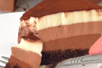 Slice of multi-layer chocolate and vanilla mousse cake with cocoa dust on top, being cut with a fork on a plate.