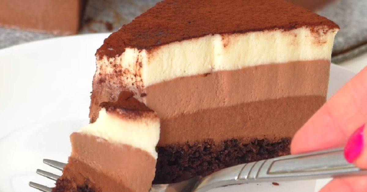 Slice of multi-layer chocolate and vanilla mousse cake with cocoa dust on top, being cut with a fork on a plate.