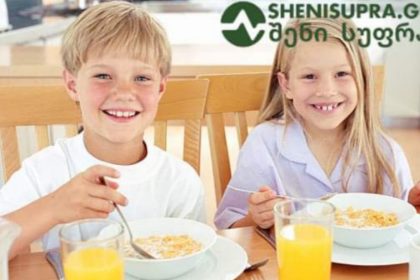 Two smiling children, a boy and a girl, eat cereal at a wooden table with bowls and glasses of orange juice.