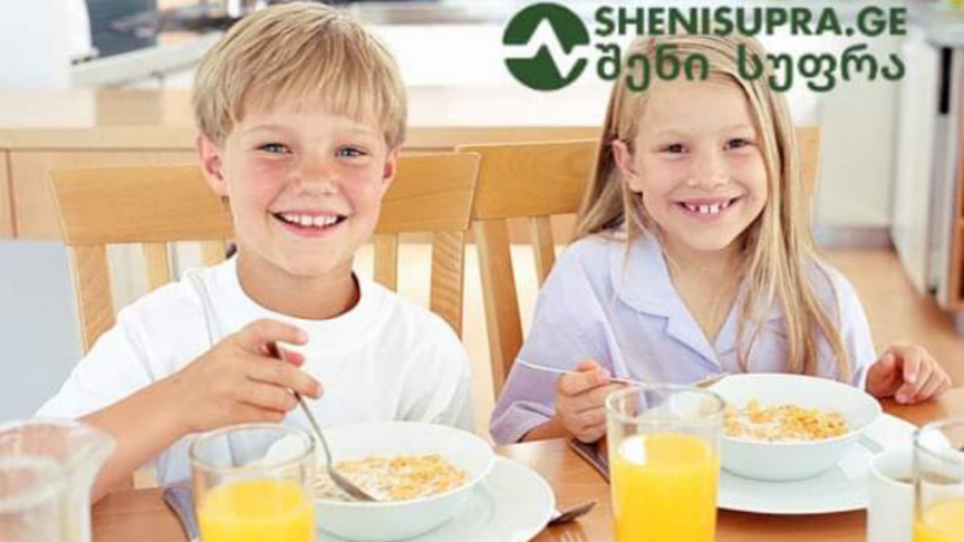 Two smiling children, a boy and a girl, eat cereal at a wooden table with bowls and glasses of orange juice.