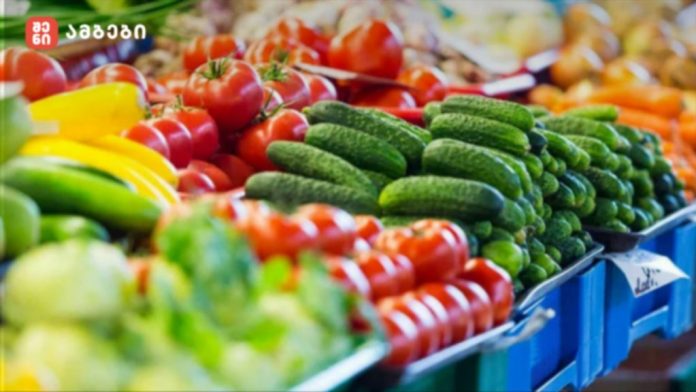 sheniambebi-produqtebi-1024x576 Assorted fresh vegetables—cucumbers, tomatoes, and peppers—arranged in crates at a market stall.