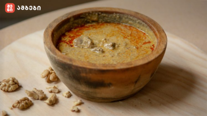 Rustic clay bowl of creamy walnut soup with paprika oil, on a wooden board with walnut pieces nearby.