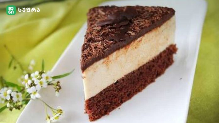 shenisupra-ahokoladis-torti Slice of chocolate–vanilla layered cake on a white plate with a green background and small white flowers nearby