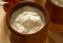 როგორ მოვამზადოთ არაჟანი Creamy yogurt served in a brown ceramic pot on a wooden table, with tomatoes visible in the background.
