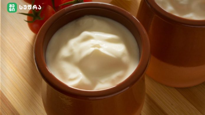 Creamy yogurt served in a brown ceramic pot on a wooden table, with tomatoes visible in the background.