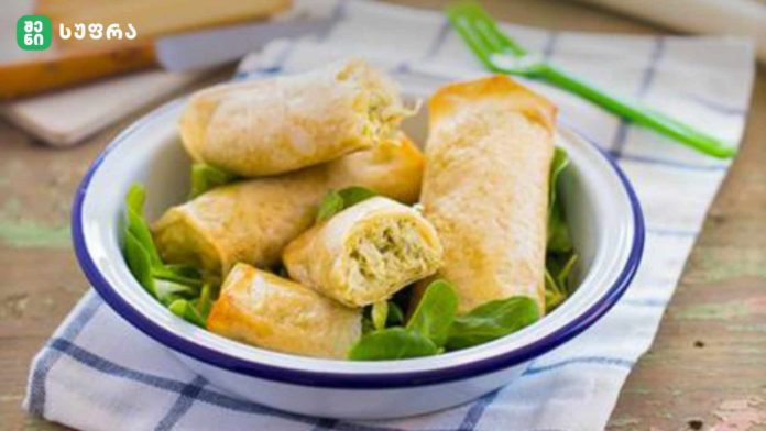 White bowl with blue rim filled with golden spring rolls atop green lettuce on a checkered cloth.