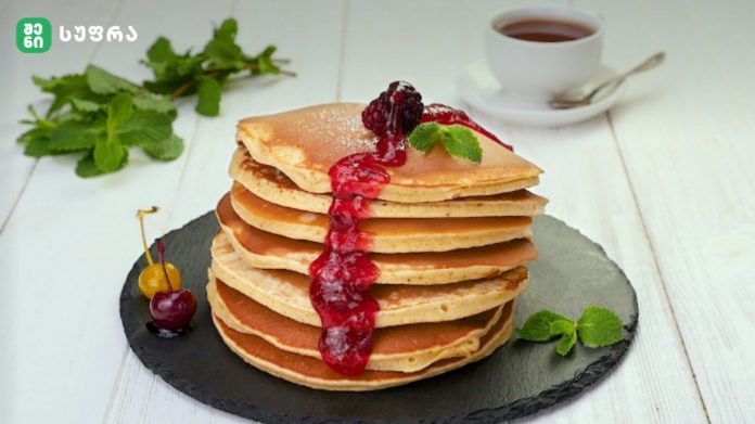 shenisupra-blinebi-3 Stack of golden pancakes stacked high, drenched in red berry sauce with mint garnish on a dark slate board; tea cup in the background.