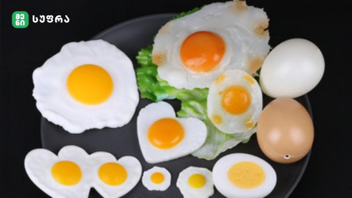 Assorted fried eggs in different shapes and sizes arranged on a black plate with greens and a logo in the corner.