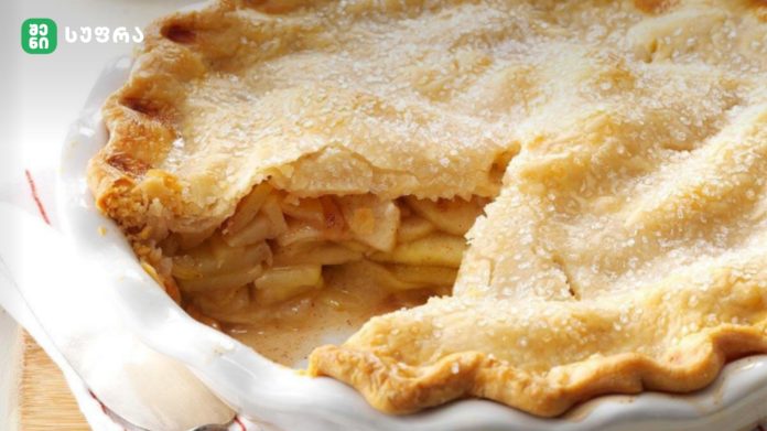 shenisupra-vashli Brand logo in the upper-left corner over a close-up of a flaky, sugar-dusted apple pie in a white dish. Typical baked filling visible.
