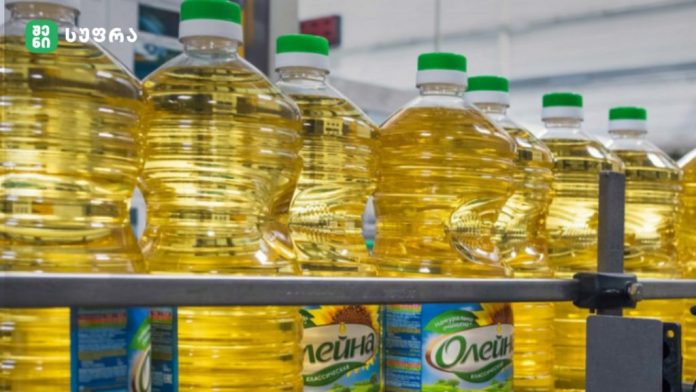 Rows of large transparent cooking oil bottles with green caps on a metal conveyor in a bottling facility.