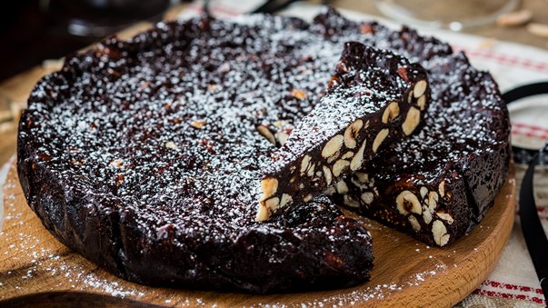 sienuri panporte Dense chocolate loaf cake studded with almonds, sliced to reveal a rich interior, lightly dusted with powdered sugar on a wooden board.