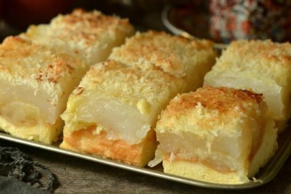 Tray of stacked dessert squares with a pale yellow cake base, an orange filling, a clear gelatin center, and a coconut crumb topping on a metal tray