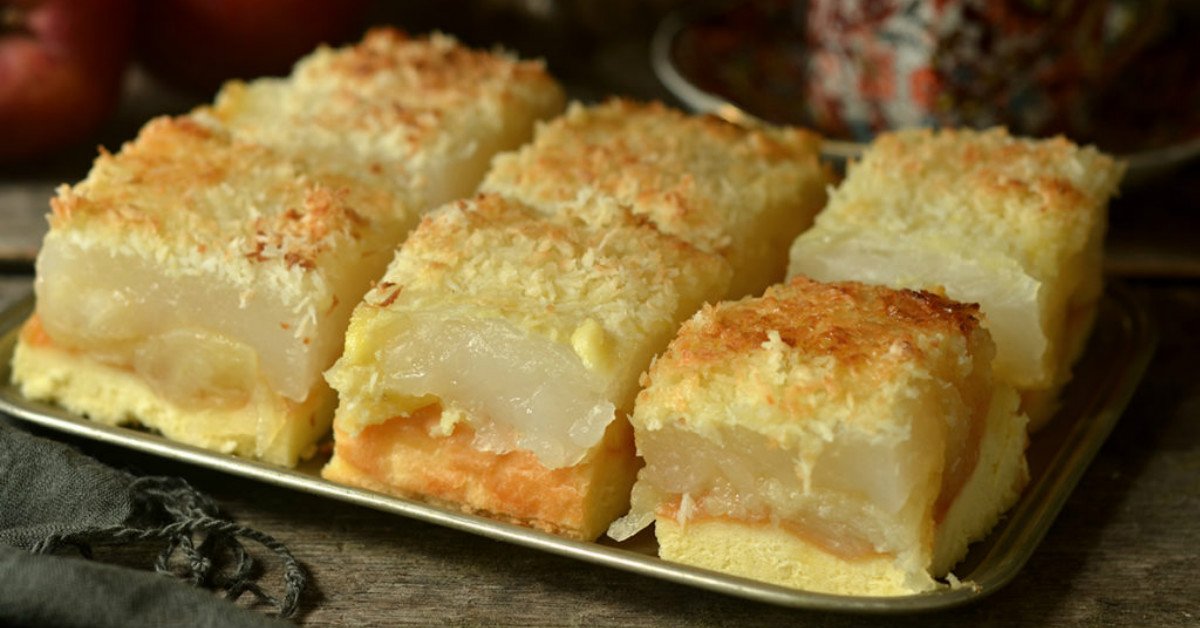 Tray of stacked dessert squares with a pale yellow cake base, an orange filling, a clear gelatin center, and a coconut crumb topping on a metal tray